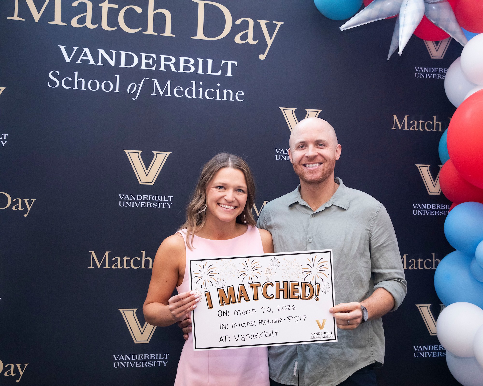 A physician-scientist couple holds their match day card front of a festive backdrop.