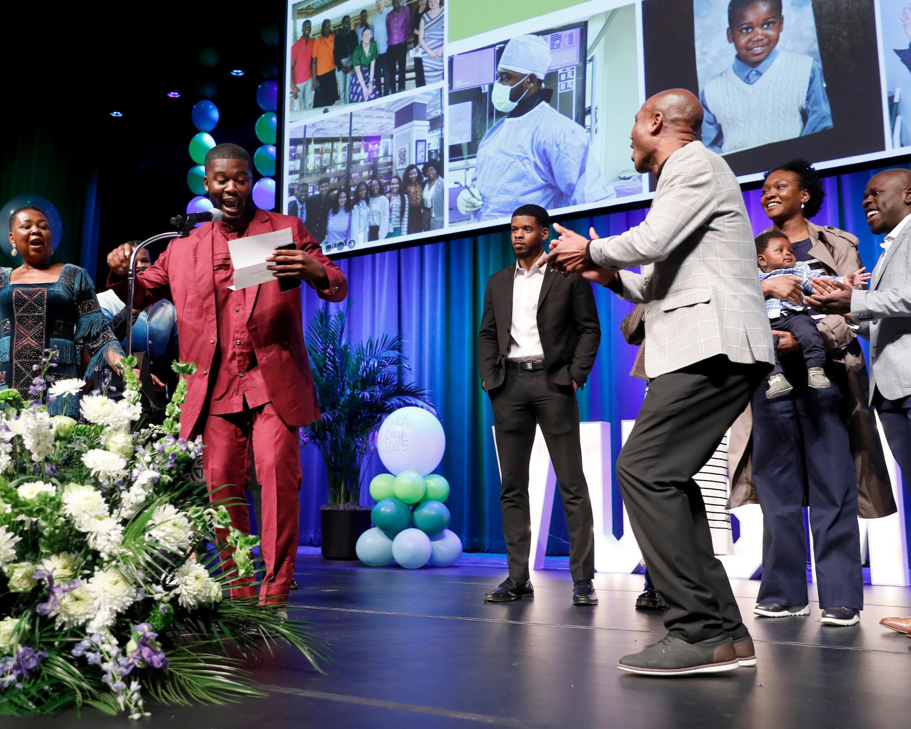 A Black man in a red suit celebrates with his family onstage at a medical residency match day event.
