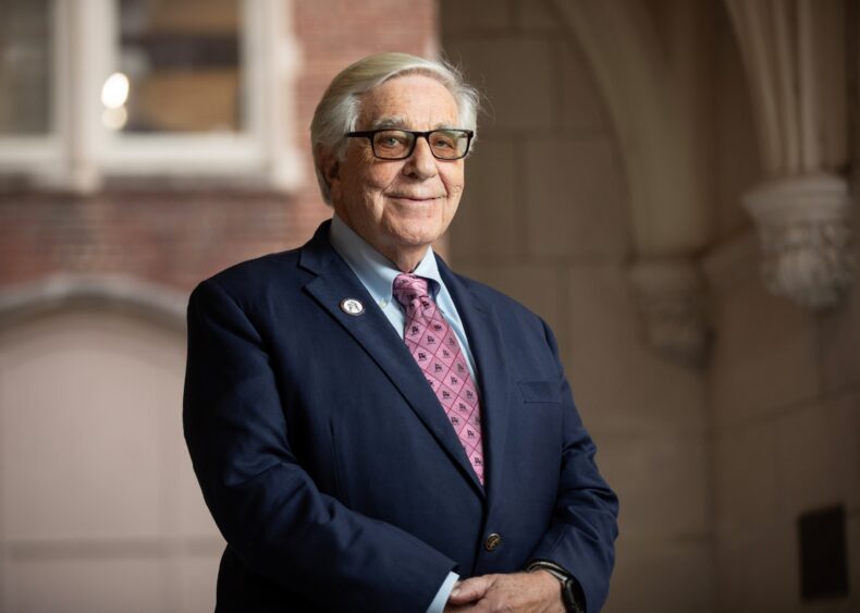Dr. Billy Hudson, an older white male wearing a business suit and glasses, poses in front of a brick and stone academic building.