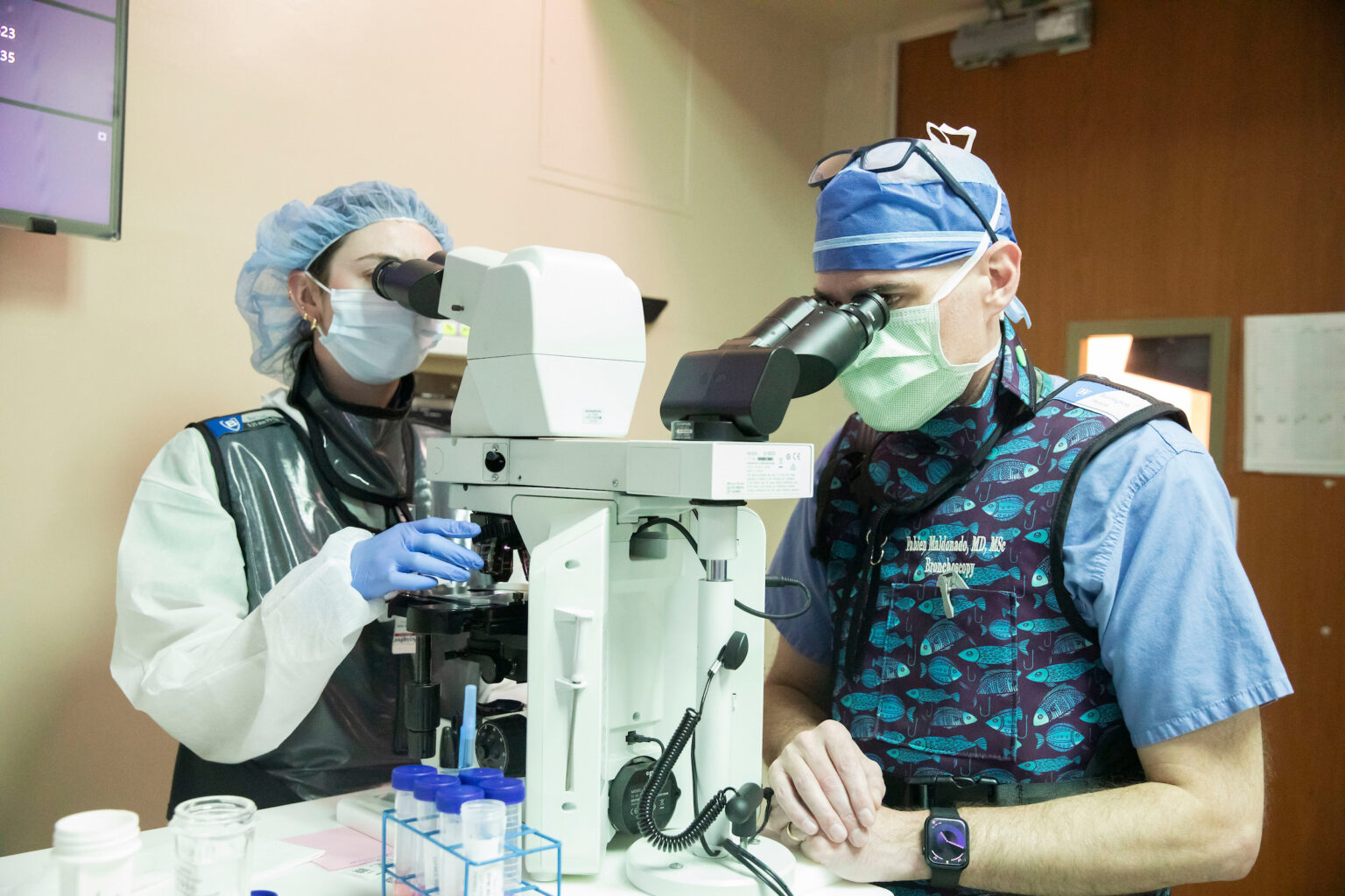Two medical professionals in scrubs and surgical masks look through microscopes in a hospital room.