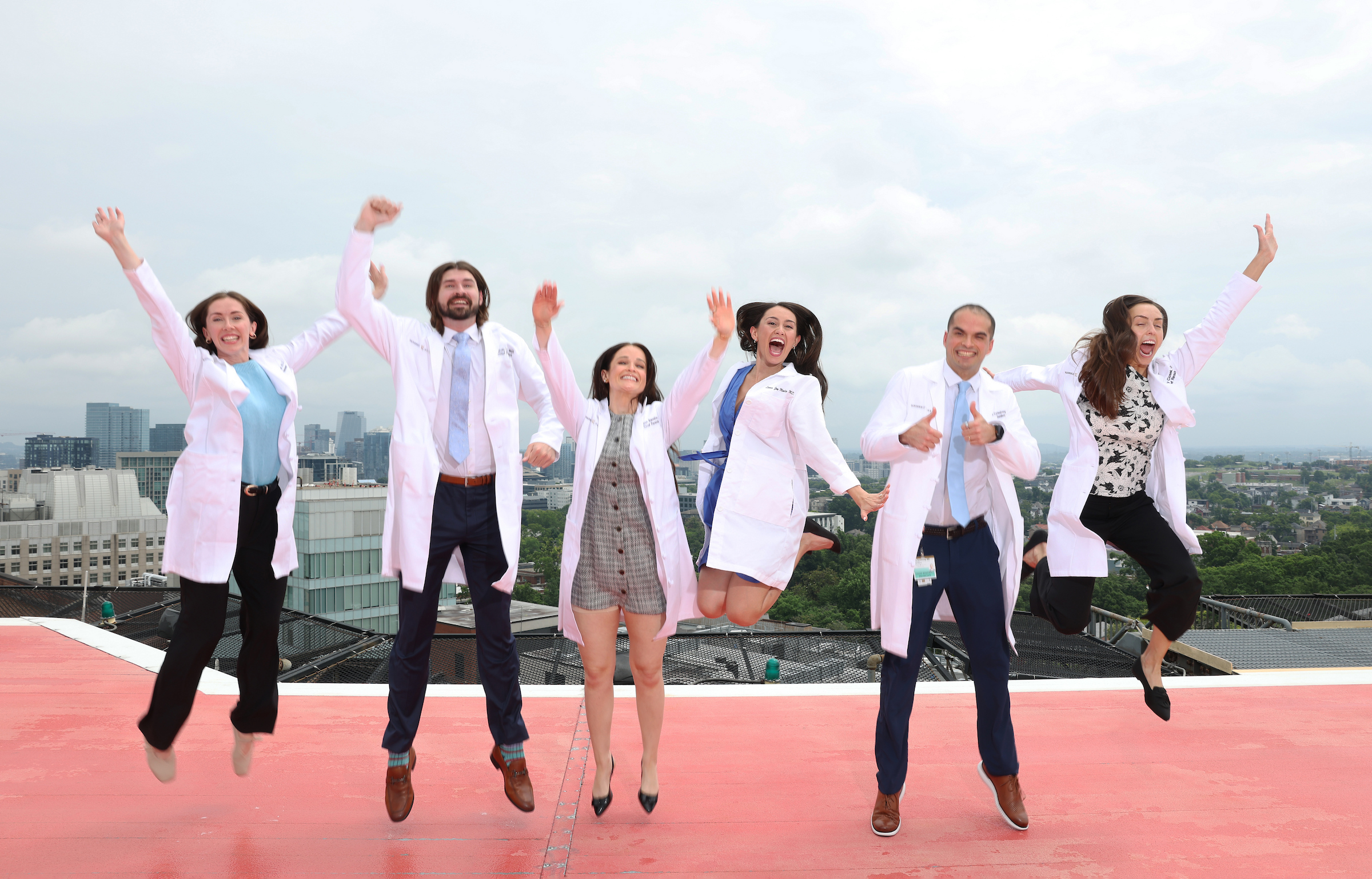Six chief residents in white coats jump in the air on the hospital roof helipad.