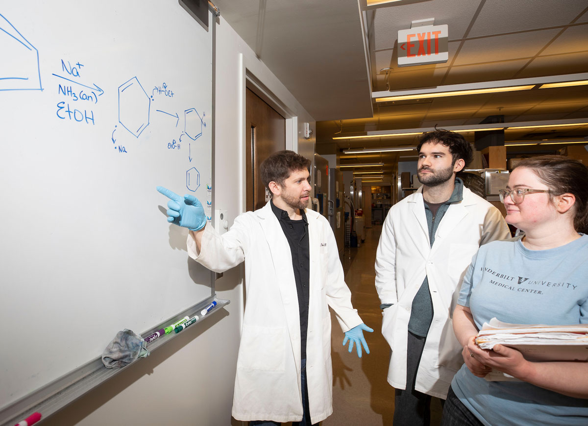 A male researcher points to a formula on a white board as two students look on.