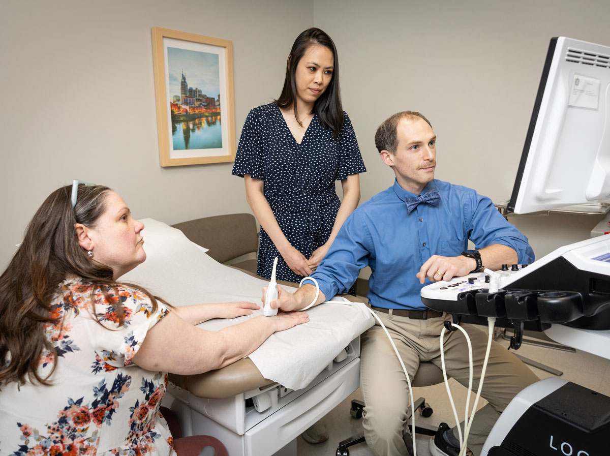 A male doctor examines a patient's hand with an ultrasound while a female doctor looks on.