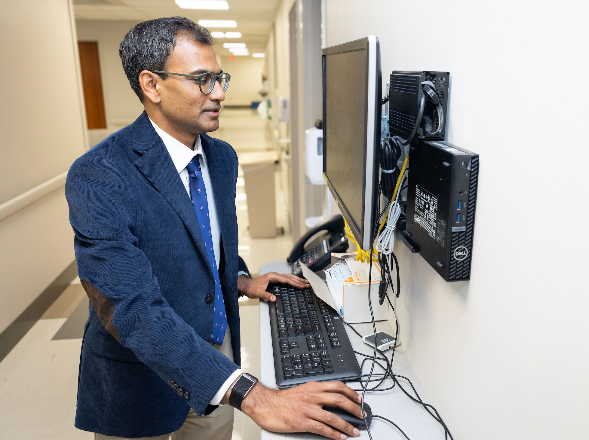 A male doctor works at a computer station in a hospital hallway.