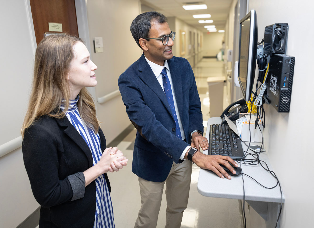 A male rheumvaology fellowship mentor looks at a computer screen in a hospital hallway as a female researcher looks on.