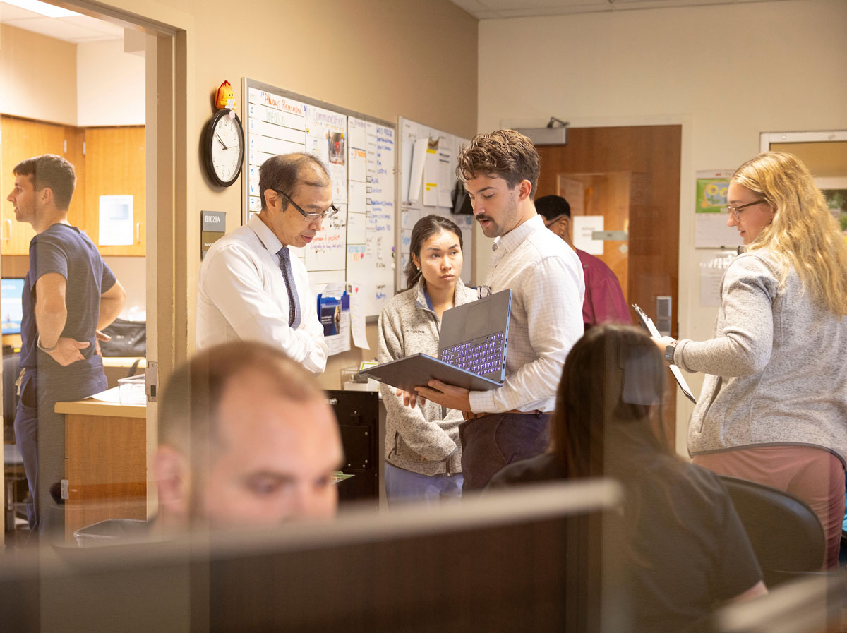 Several physicians talk while looking at a laptop while others work around them at a hospital work station.