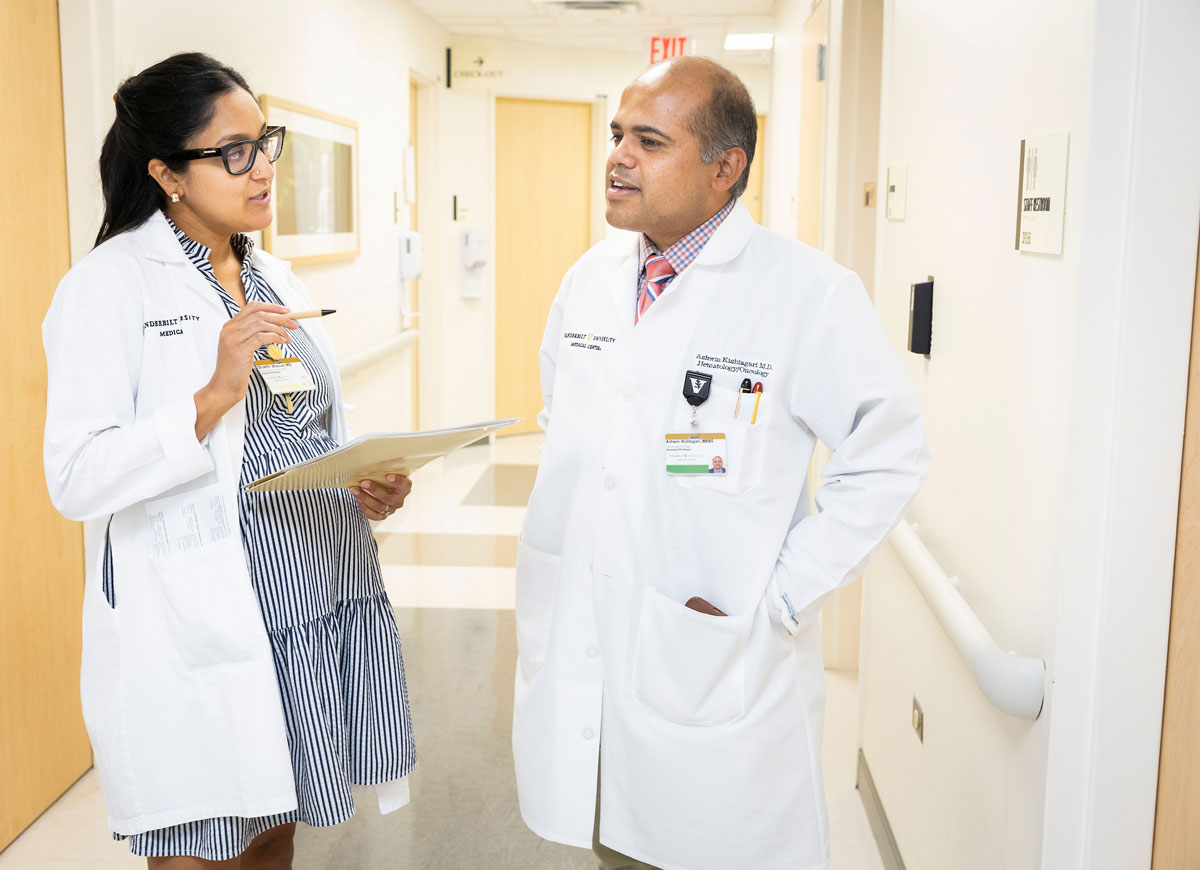 A male and female doctor in white coats chat in a hospital hallway.