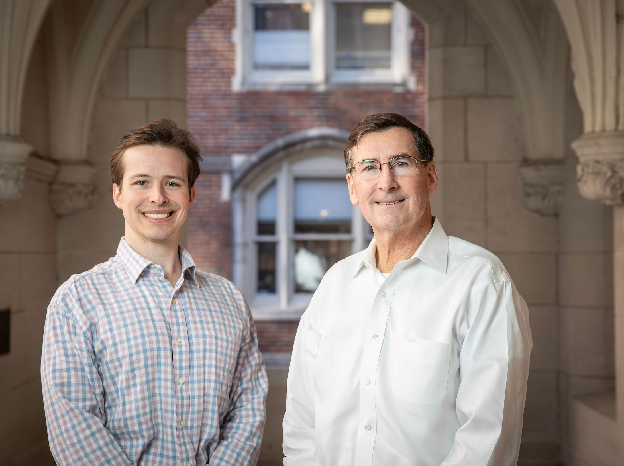 Two white male researchers pose in front of a stone and brick academic building.