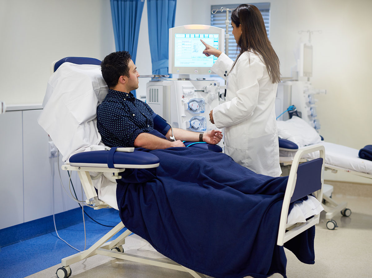 A female doctor sits on the bedside of a male patient to assist him with a dialysis treatment.