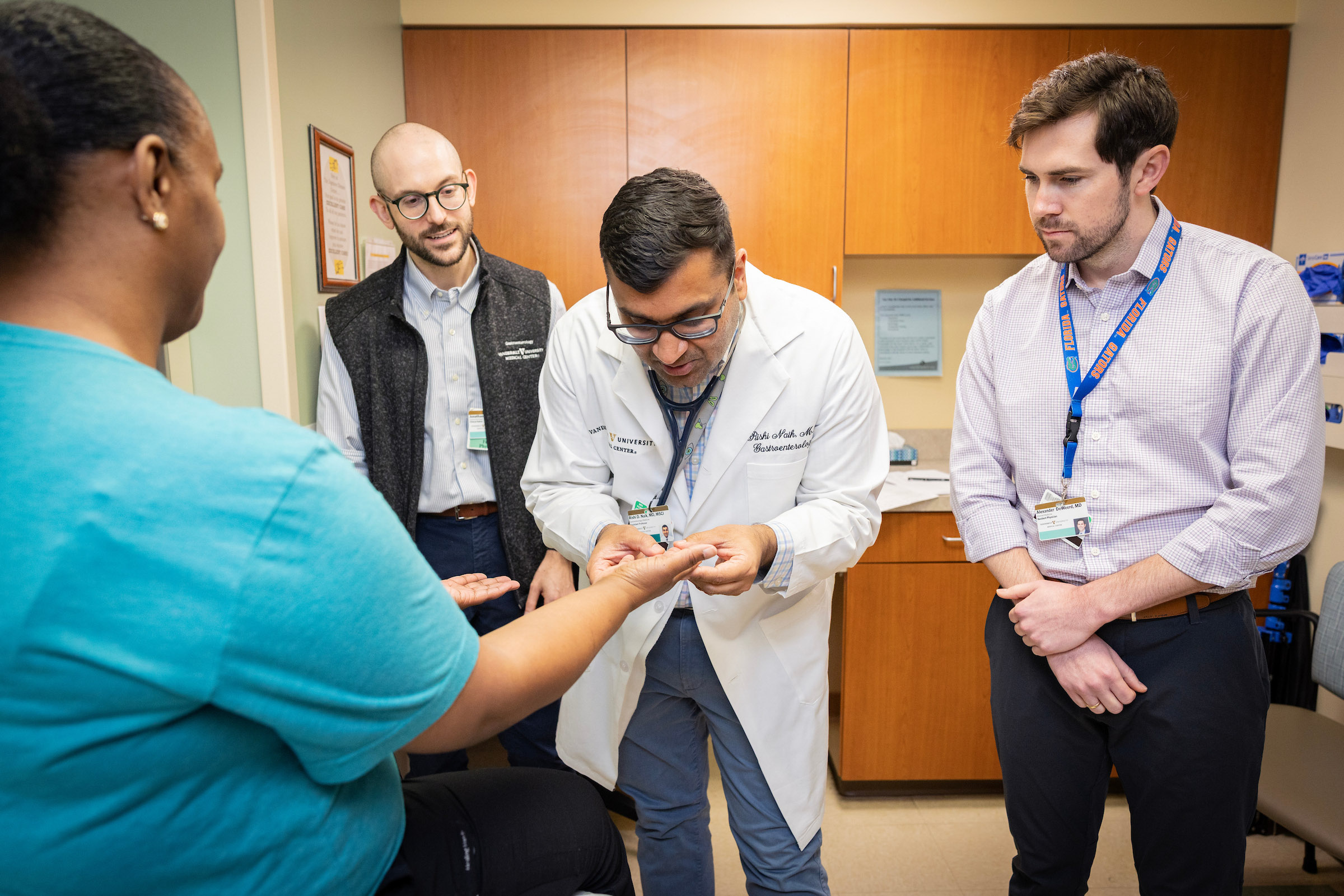 A male doctor examines a female patient's hand while two other male doctors observe.