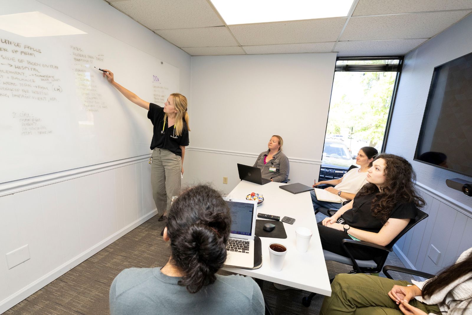 A physician instructor points to a whiteboard while a small group of female students looks on.