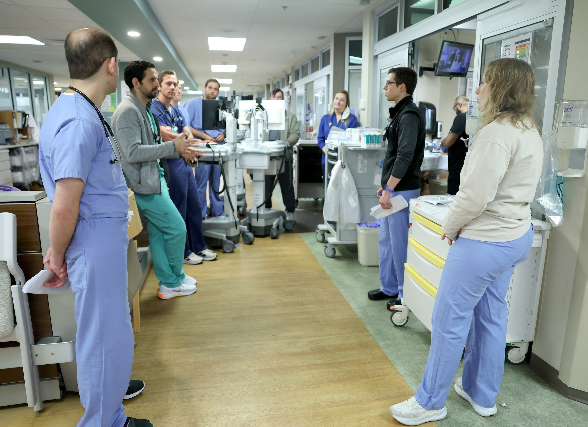 A group of doctors in blue scrubs talk in a hospital hallway.