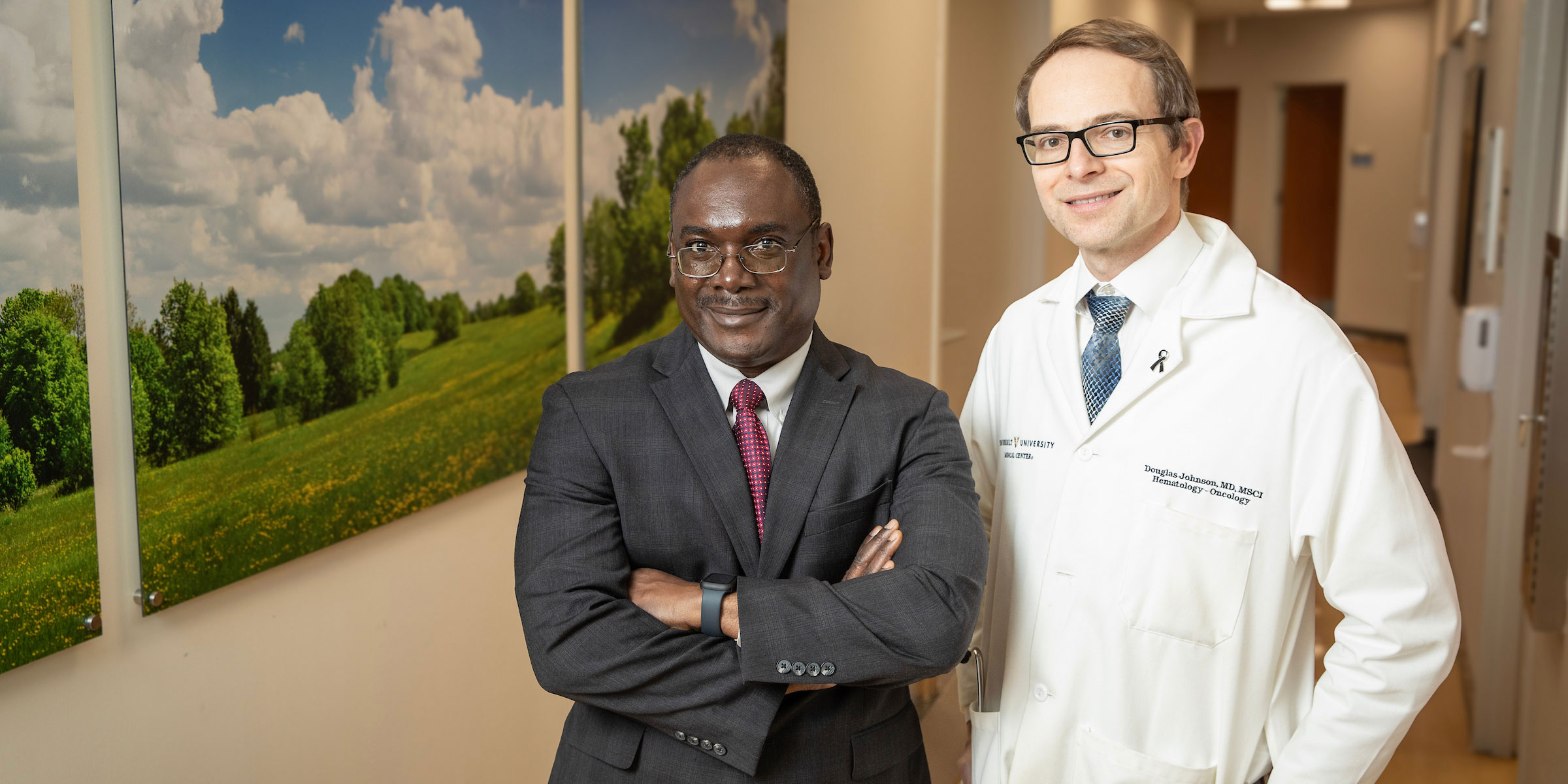 One Black and one white male physician pose for a photo in a hospital hallway.