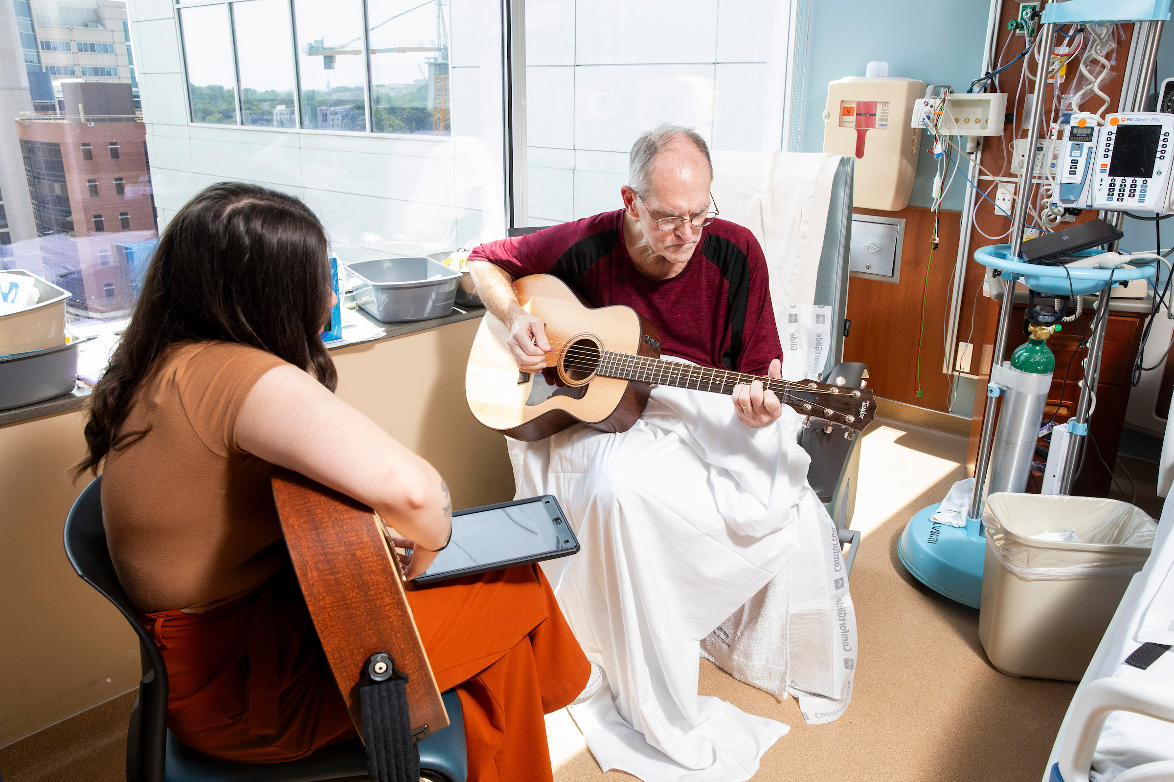 A young female music therapist plays guitar with an older male patient in his room.