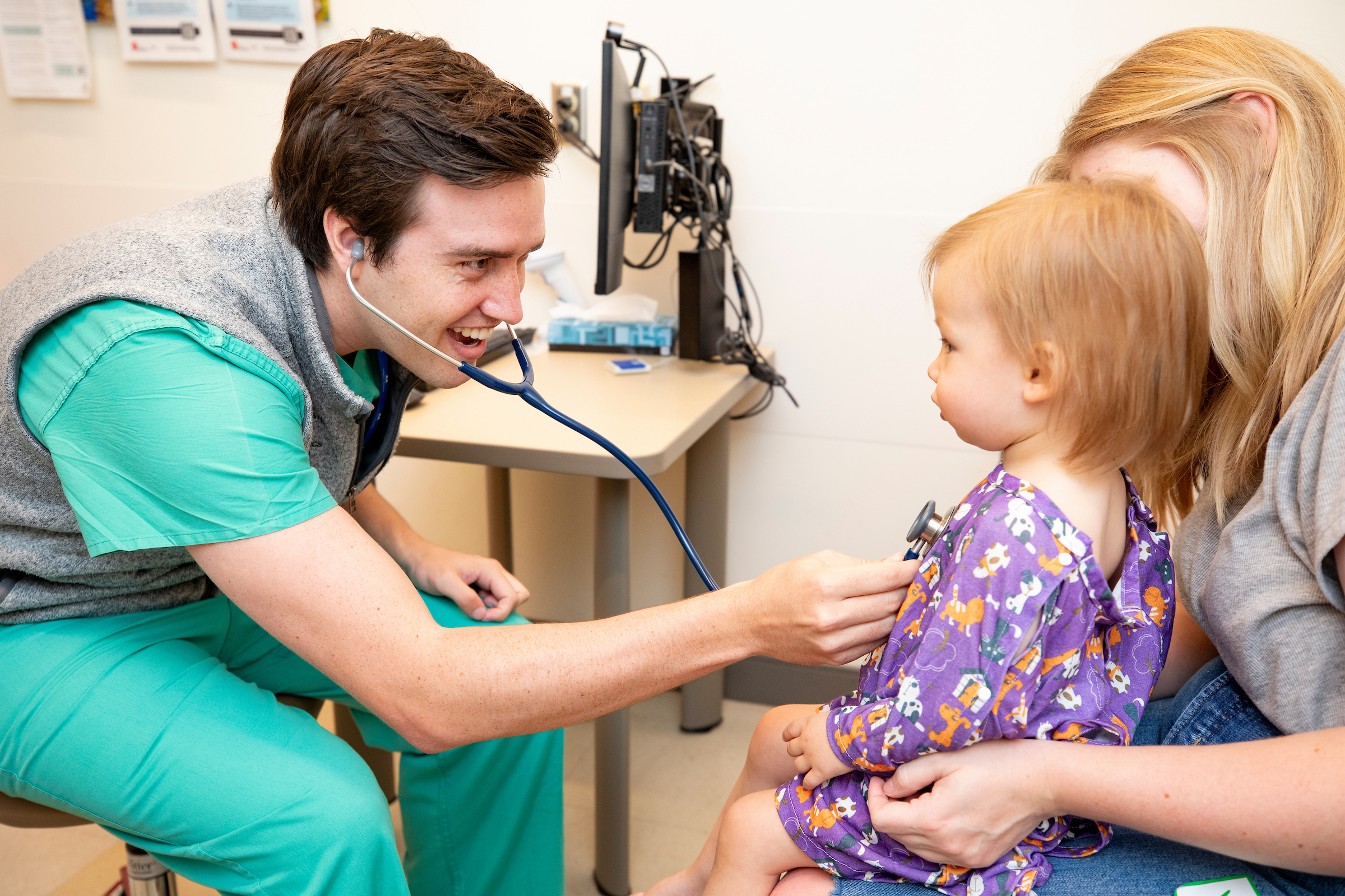 A male resident laughs with a toddler patient and her mother in an exam room.