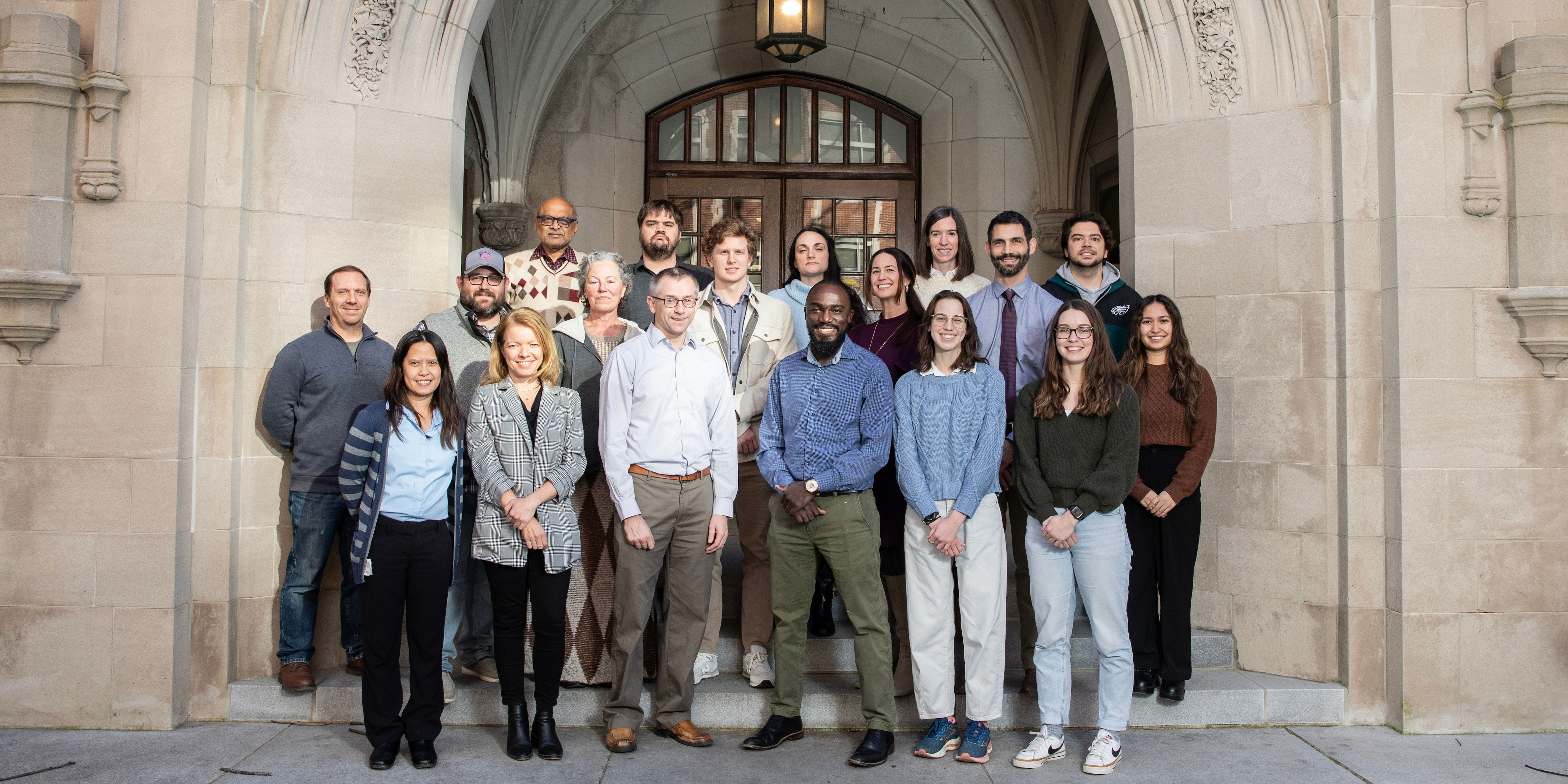 About 15 research lab members pose in front of a stone and brick academic building.