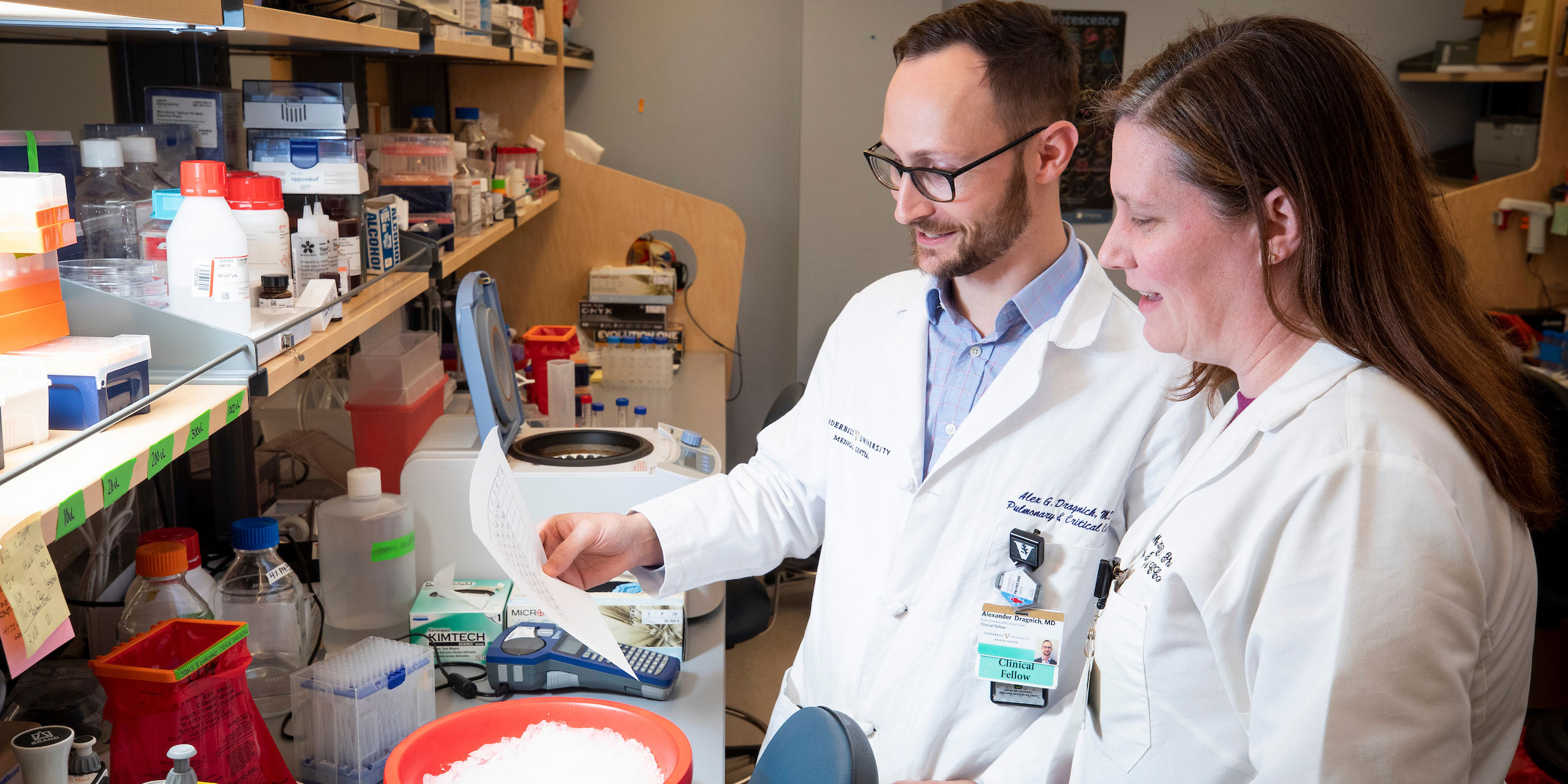 One male and one female research scientist in white lab coats look at look at materials in the lab.