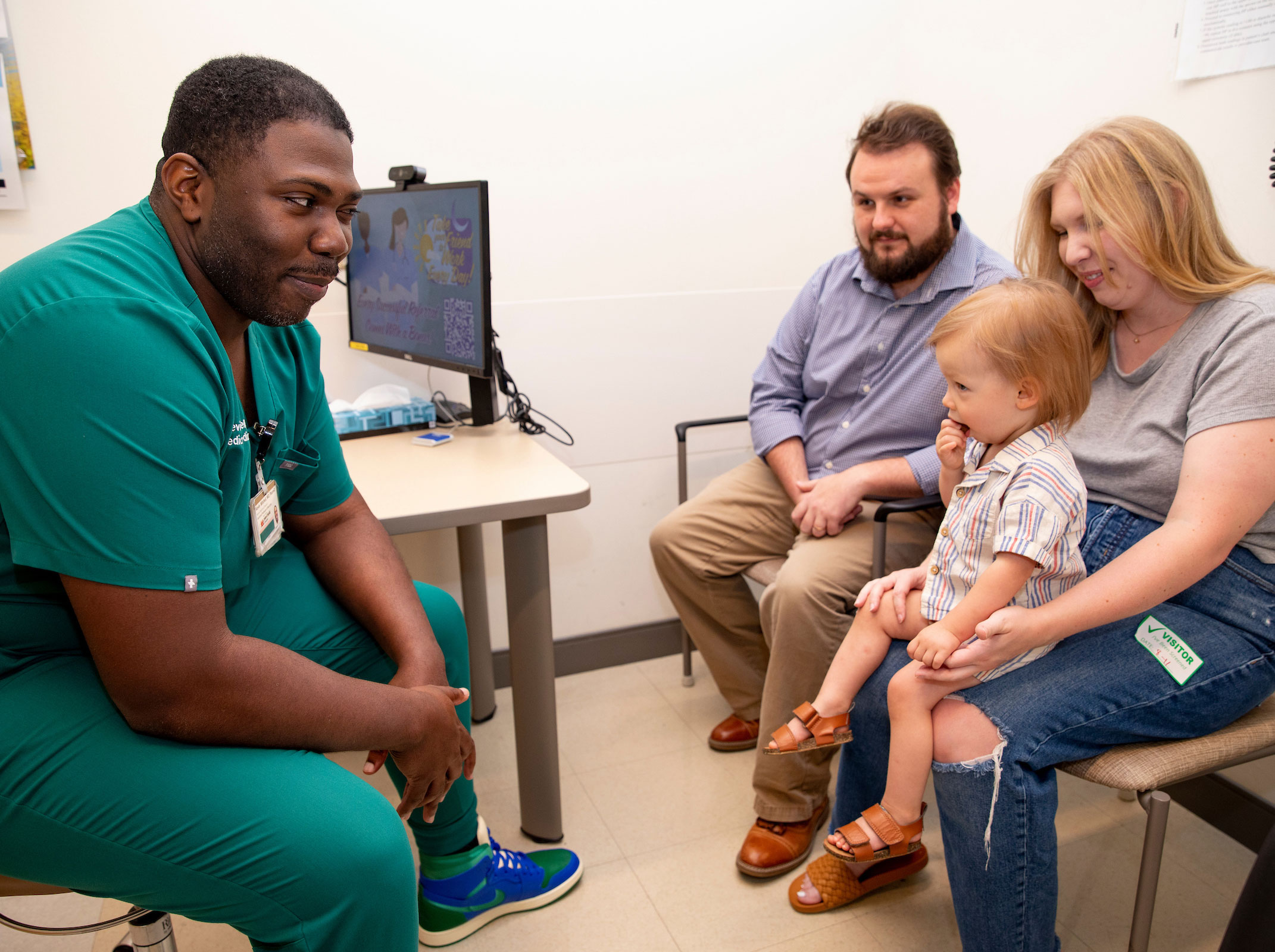A Black male doctor in scrubs talks with parents and their young child in an exam room.
