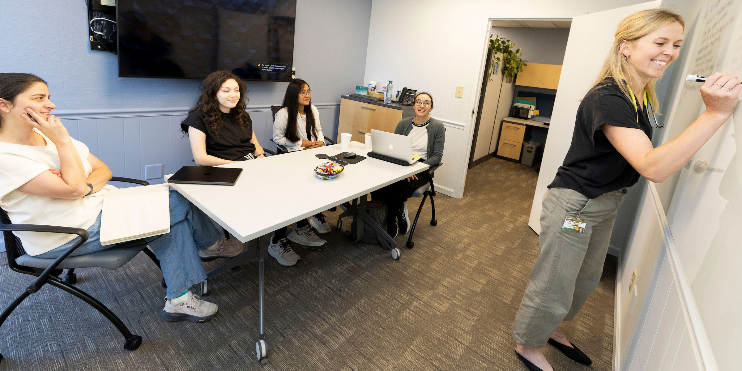A female physician draws on a white board in a small classroom with four geriatric fellows listening.