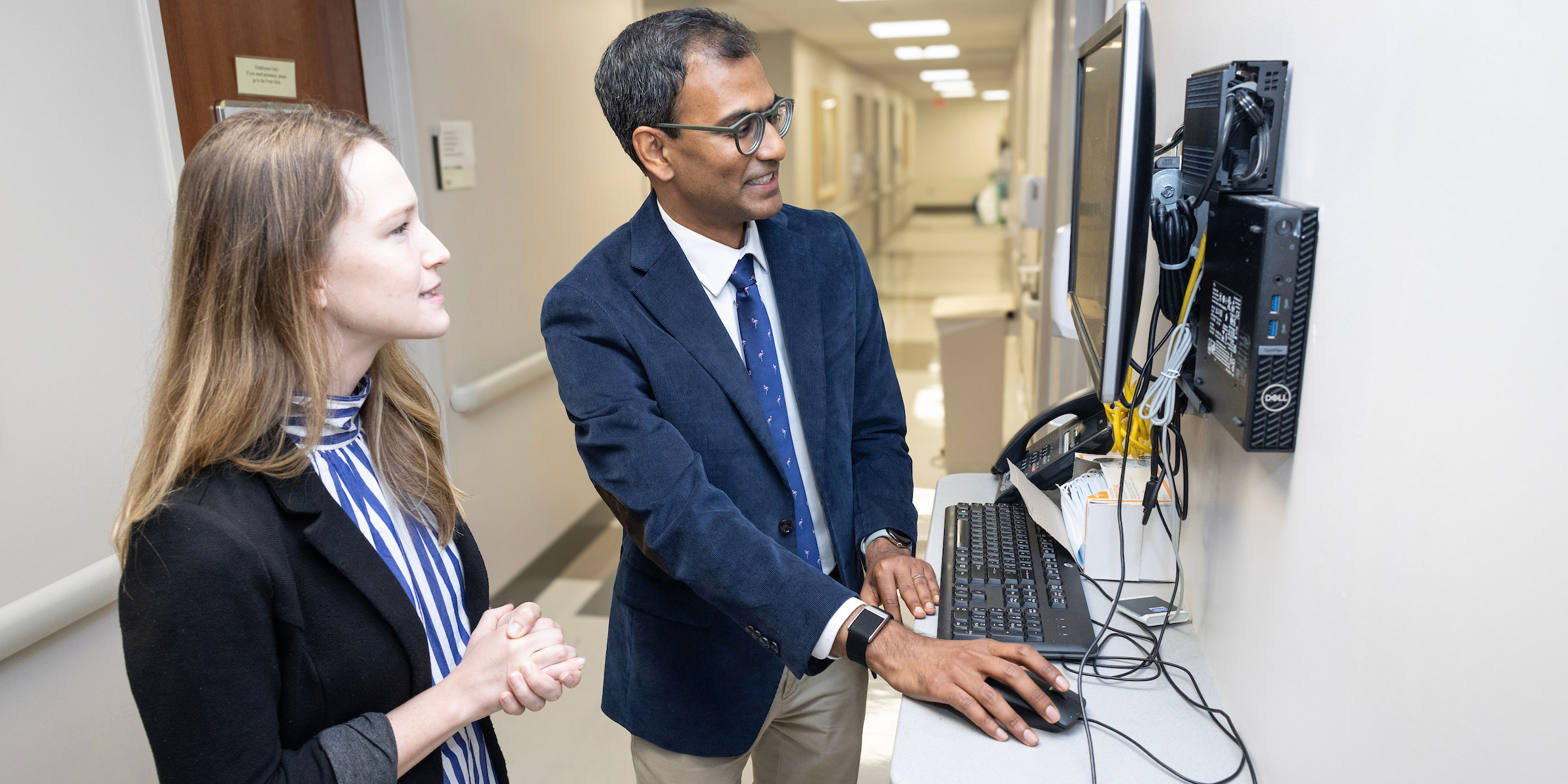 A male and female physician look at a computer screen in a hospital hallway.