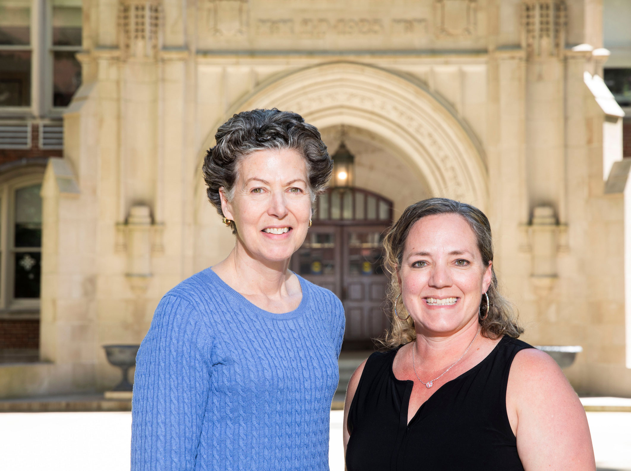 Two female research scientists pose in front of a stone and brick academic building.