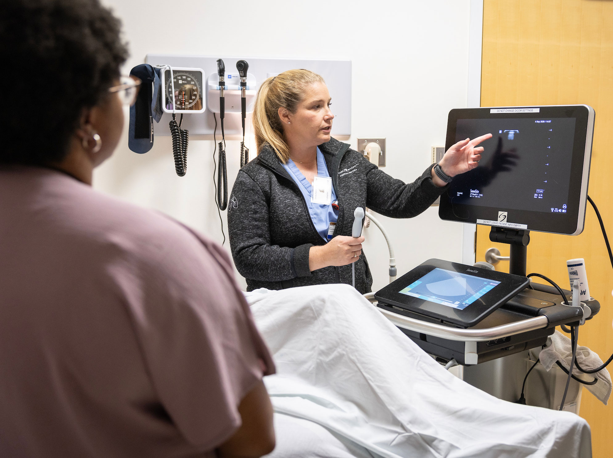 A nurse demonstrates an ultrasound on a patient volunteer while 1 other nurse looks on.