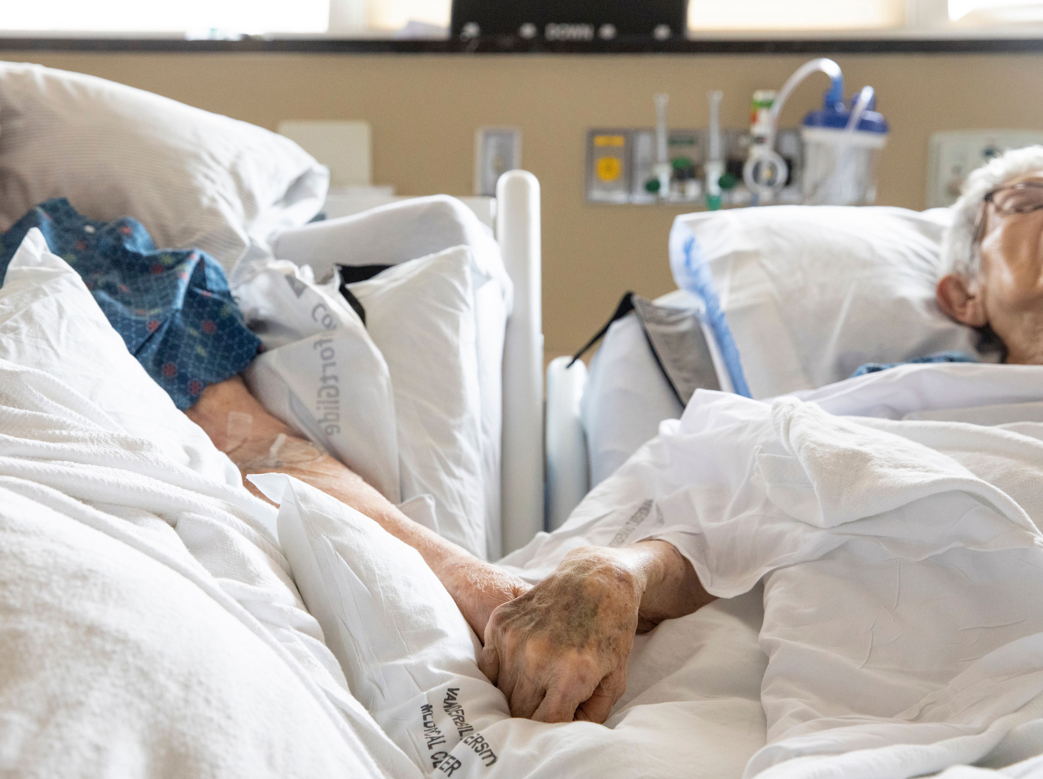 Two elderly patients in hospital beds hold hands.