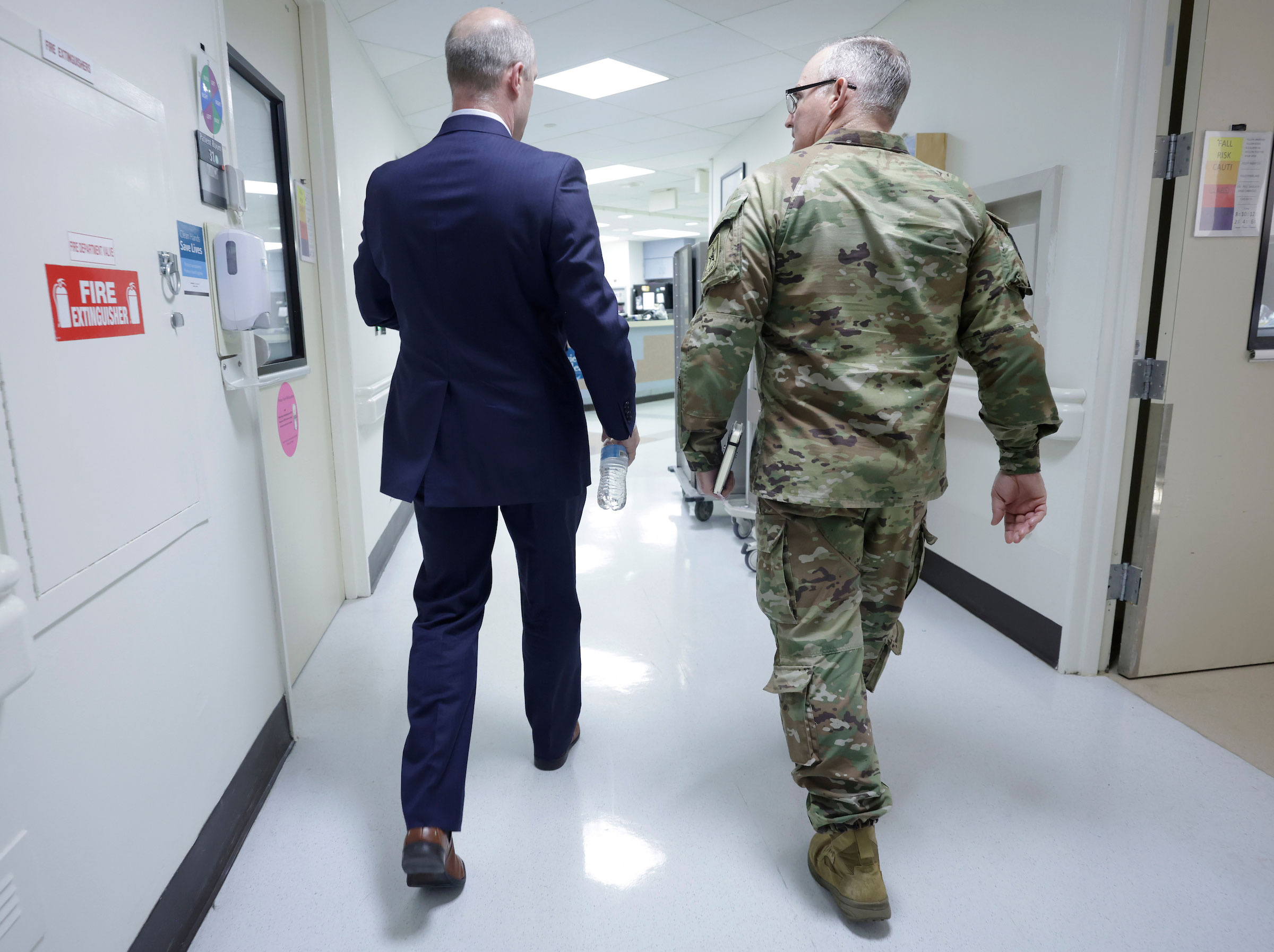 A man in green Army fatigues walks in a hospital hallway with a man in a dark blue suit.