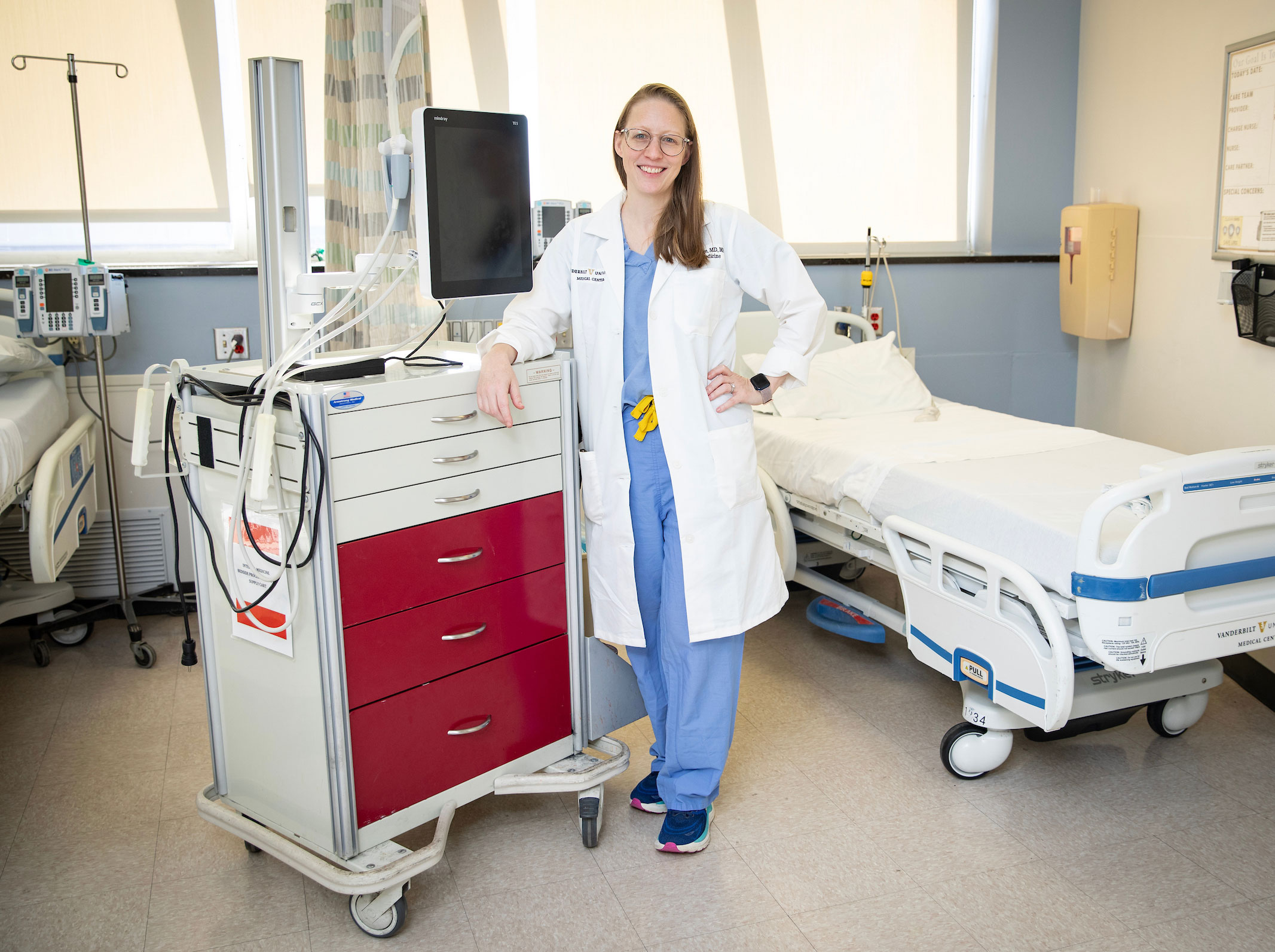 A female doctor poses in an empty hospital patient room with an equipment cart.