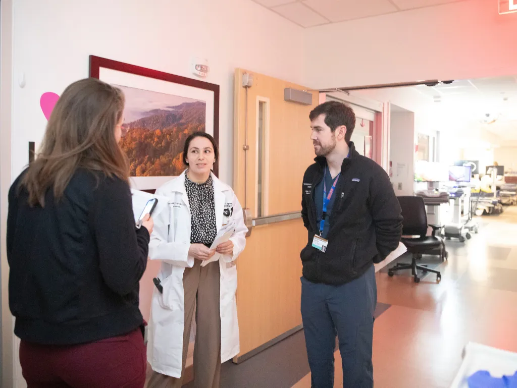 A female physician in a white lab coat talks with 2 residents in a hospital hallway