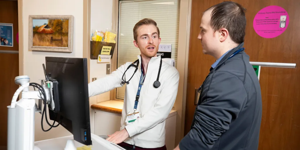 Two White male doctors look at a computer screen in a hospital hallway. 