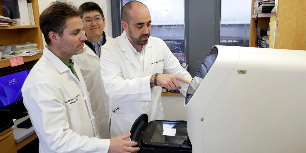 Three male research scientists look at an instrument in their lab.