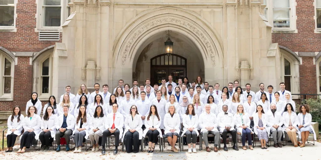 A large group of residents and faculty members in white coats pose outside a brick and stone academic building.